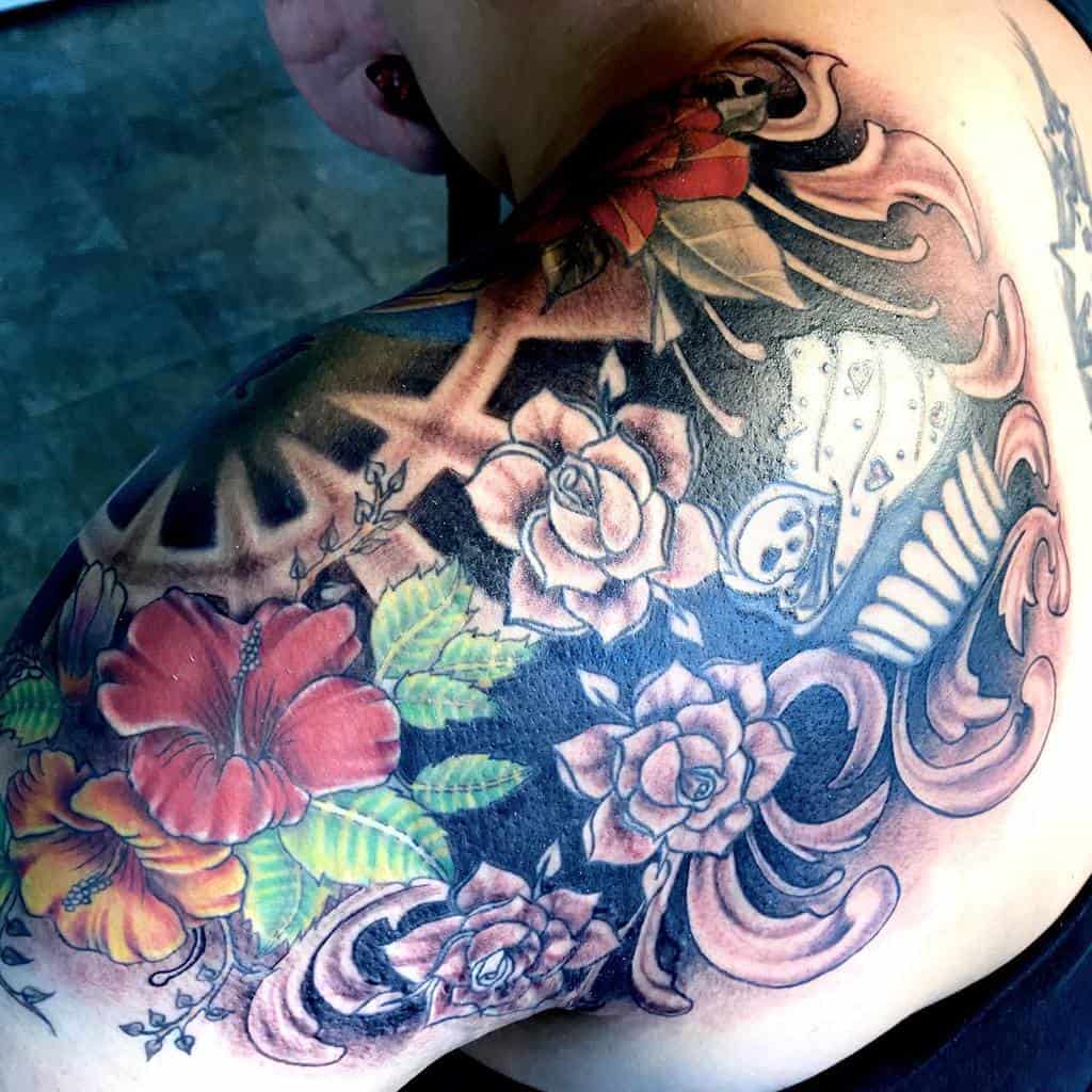 A vibrant floral tattoo artfully adorns a person's shoulder, showcasing roses and hibiscus elegantly intertwined with intricate gears and patterns