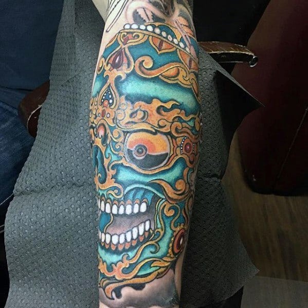 Vibrant Tibetan skull forearm tattoo with teal and orange hues, ornate patterns, and fierce expression across the lower arm.