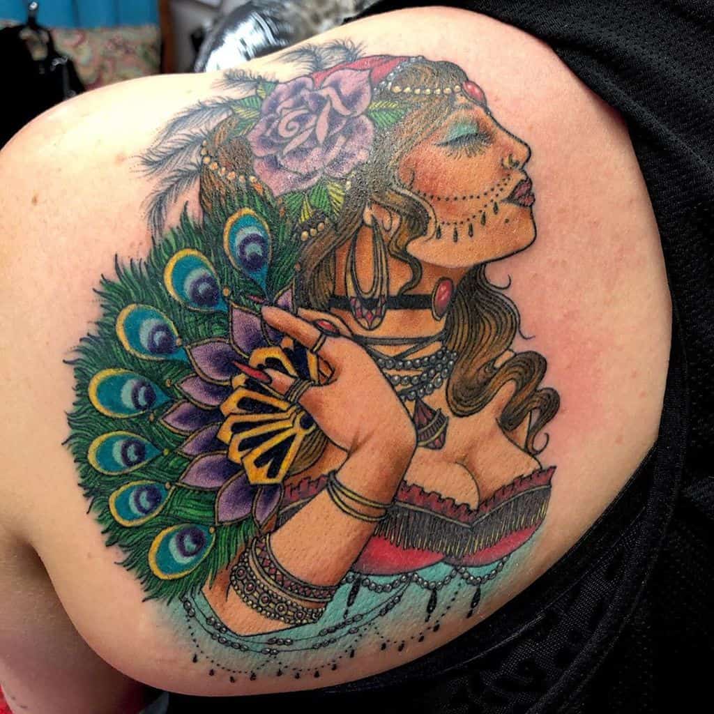 Vibrant tattoo of a gypsy woman with a peacock feather fan, intricate jewelry, and a floral headpiece on the shoulder.