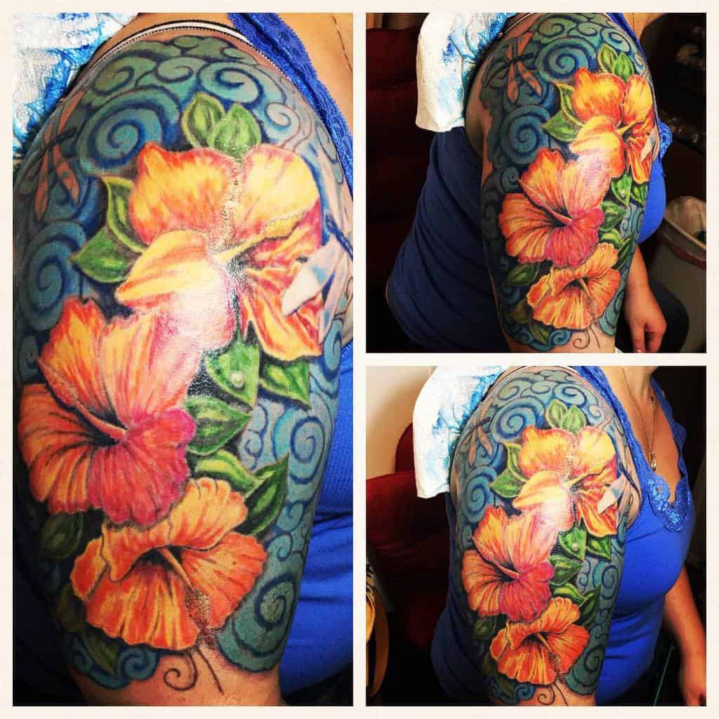 Vibrant upper arm tattoo featuring bold orange hibiscus flowers, green leaves, and swirling blue background patterns