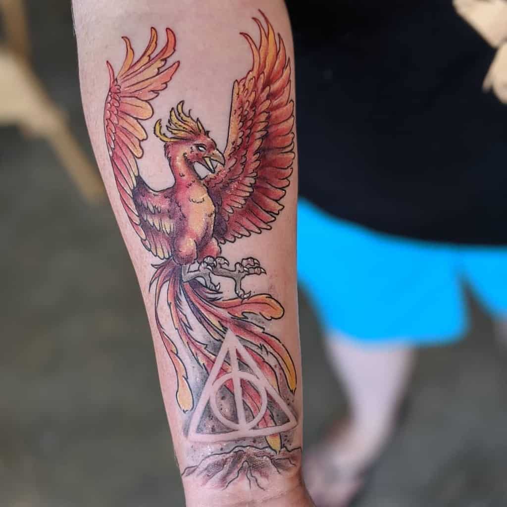 Colorful forearm tattoo featuring a fiery phoenix with outstretched wings perched on a glowing Deathly Hallows symbol atop a rocky base.