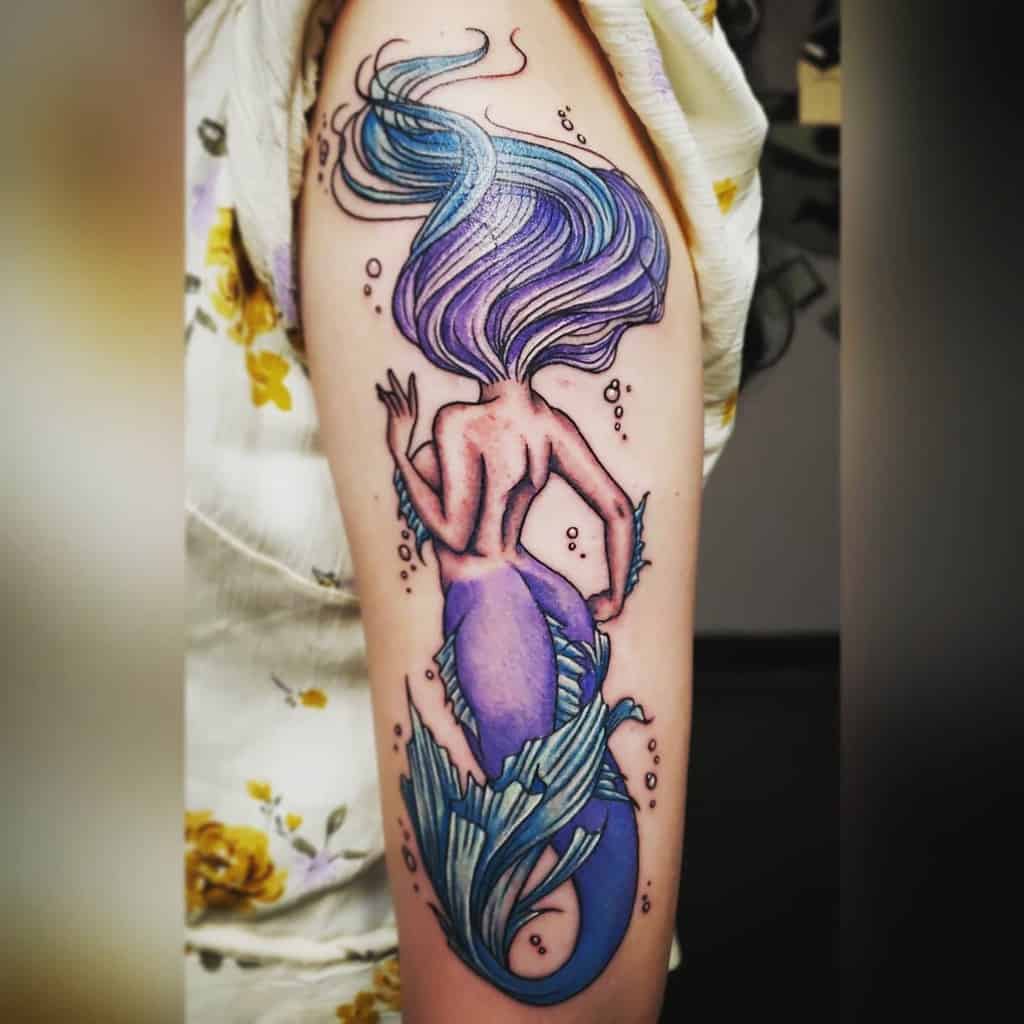 Tattoo of a mermaid with purple and blue hues on an upper arm, featuring flowing hair and a decorative tail