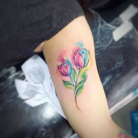 A vibrant watercolor tulip tattoo on the upper arm, featuring two colorful flowers with shades of pink, blue, and green, accented with artistic ink splashes.