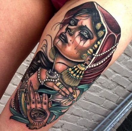 Vibrant tattoo of a gypsy woman with gold jewelry, a red headscarf, and a mystical all-seeing eye on her hand, placed on the thigh.