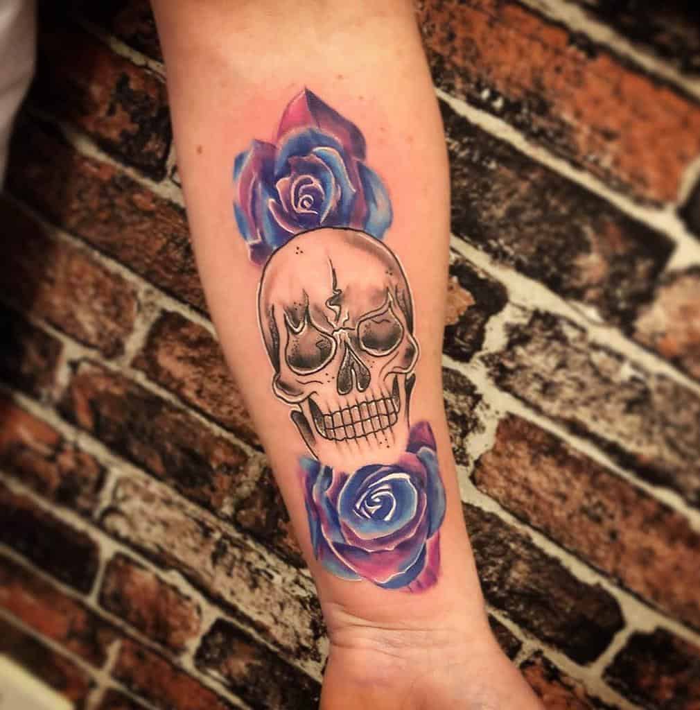 Forearm tattoo of a detailed skull framed by vibrant blue and purple roses, blending realism with watercolor-style shading.