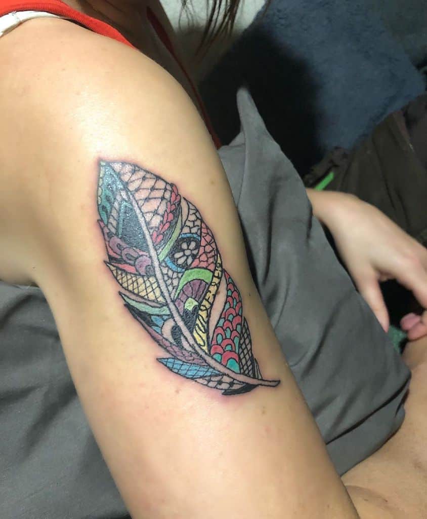 Colorful upper arm tattoo of a feather with intricate lace-like patterns, combining vibrant hues and delicate ornamental details.