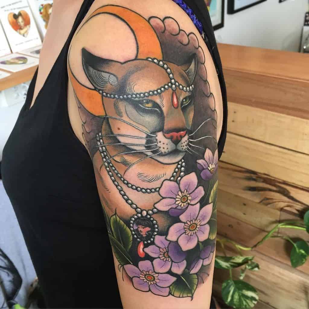 Vibrant neo-traditional lioness tattoo on the upper arm, featuring bold colors, a crescent moon, pearl necklaces, and detailed floral elements.