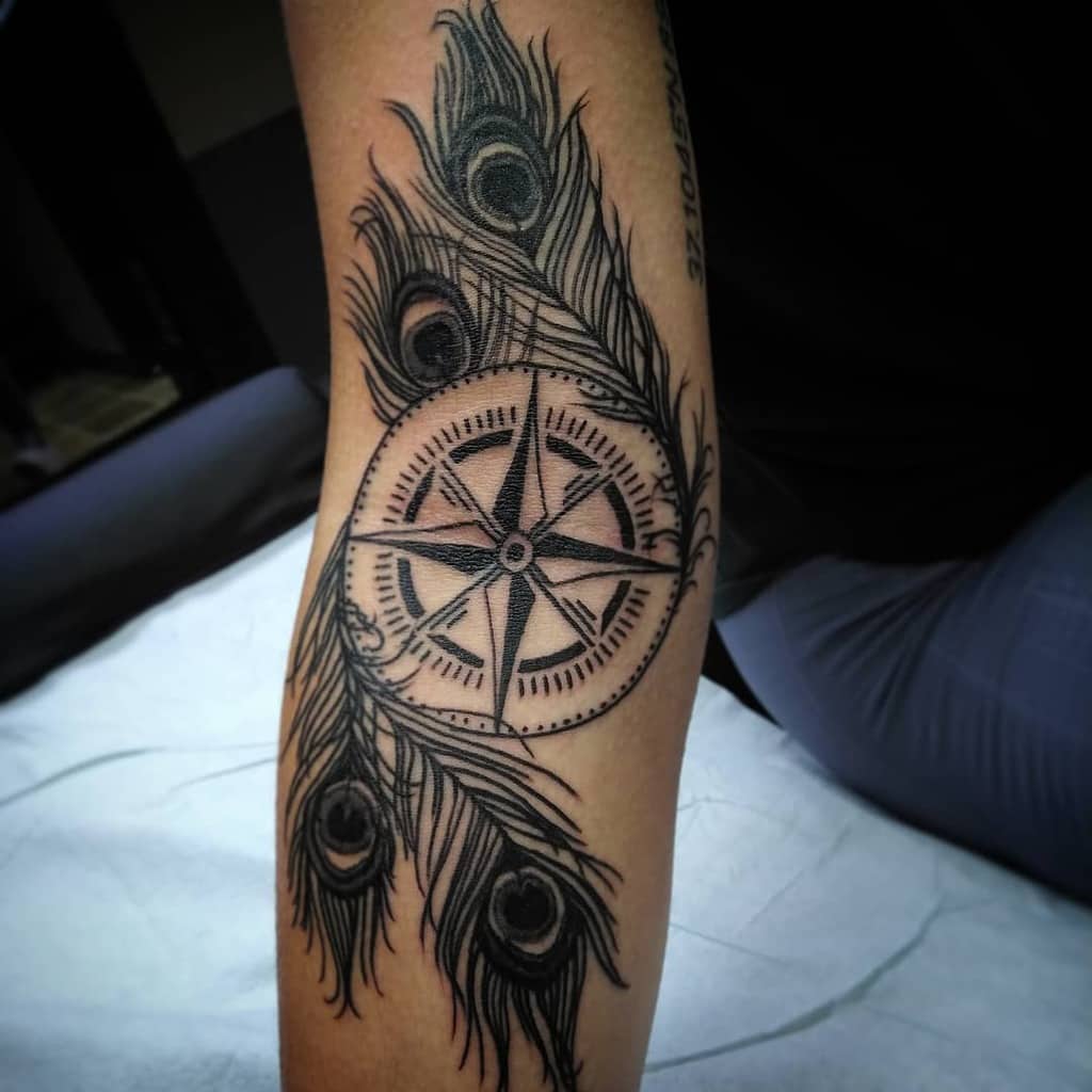 Black ink tattoo of a compass surrounded by detailed peacock feathers on the forearm, featuring fine shading and intricate linework.