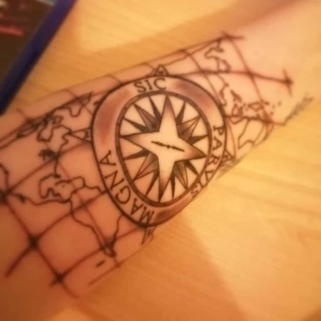 Forearm tattoo of a compass rose with "SIC PARVIS MAGNA" overlaid on a world map design