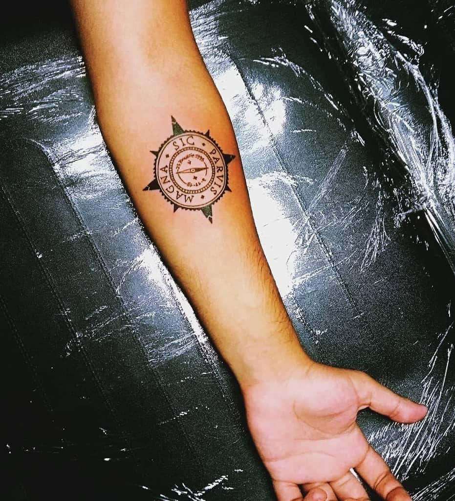 Forearm tattoo showcasing a minimalist compass design with 'Sic Parvis Magna' text in bold black ink