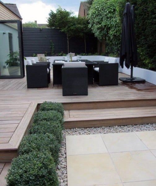 Modern wooden deck with outdoor dining set, hedges, and stone accents in a private backyard.