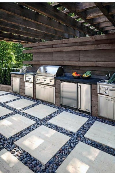 Outdoor kitchen with stainless steel appliances under a pergola, surrounded by outdoor stone and concrete pavers