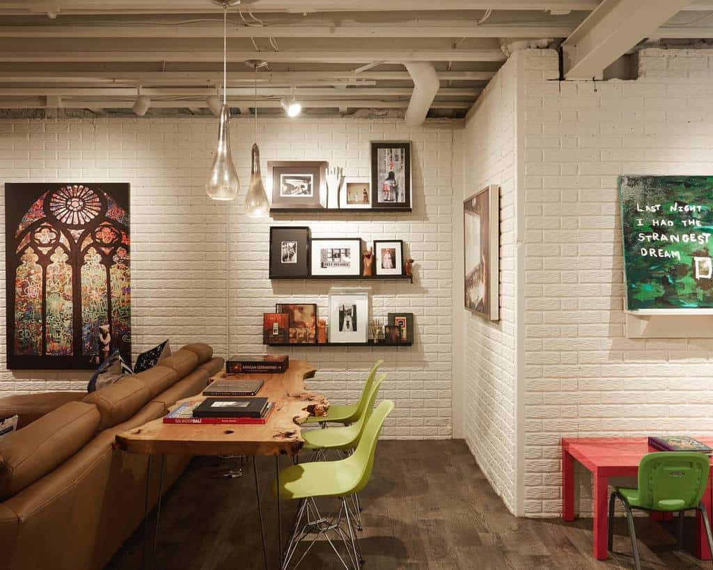 Modern basement with white brick walls, colorful artwork, and natural wood desk with green chairs.