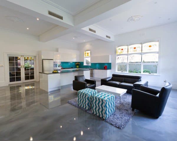 Spacious modern living room with black sofas, a blue patterned ottoman, and a bright kitchen on concrete floors