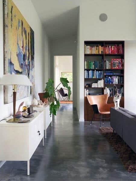 Modern living room with art, bookshelves, chairs, and a cat on a gray sofa; concrete floor and plants complete the scene