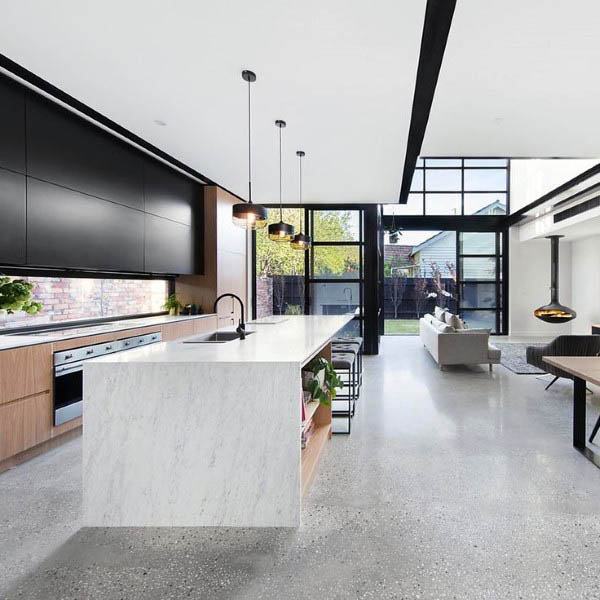 Modern kitchen with a marble island, concrete floor, hanging lights, and large windows overlooking a backyard