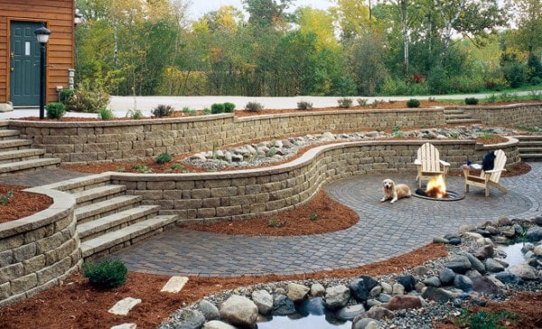 Curved stone retaining walls with stairs, a fire pit seating area, and a small rock-lined pond.