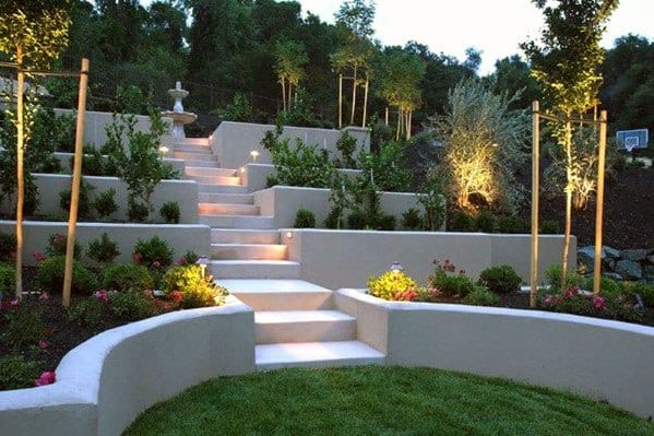 Modern sloped backyard with illuminated concrete stairs, tiered retaining walls, lush greenery, and elegant landscaping at dusk