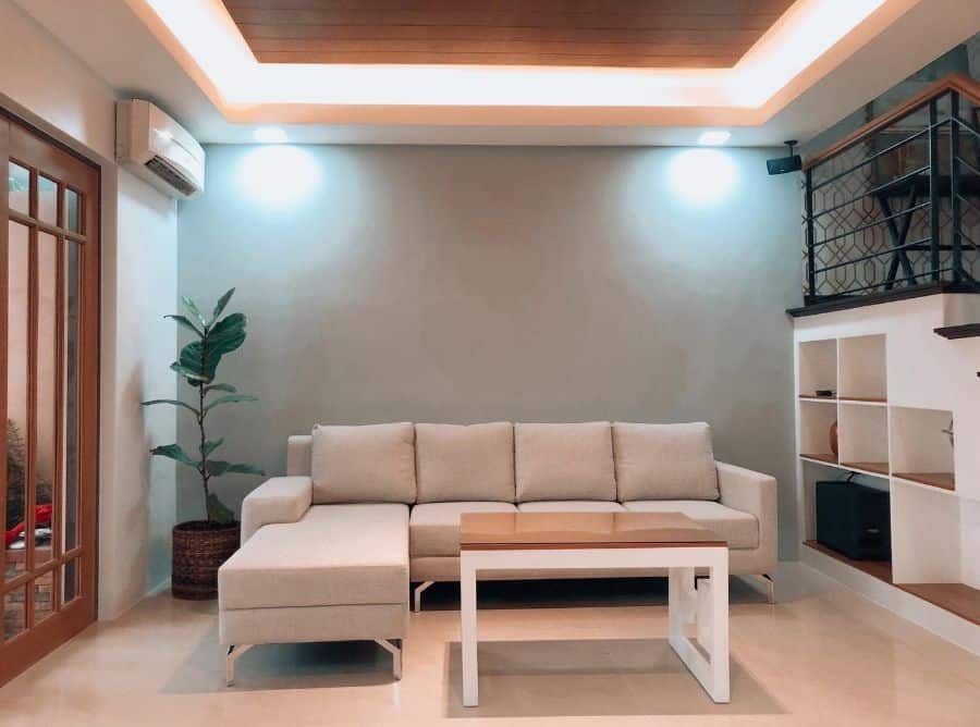 Beige sectional sofa in a minimalist living room with modern lighting, potted plant, and open shelving.