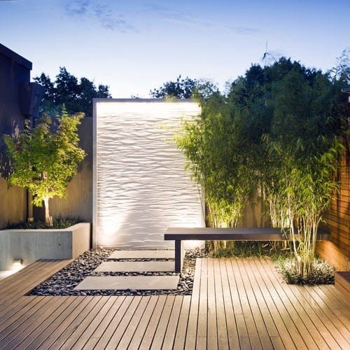 Contemporary wall fountain with textured white panel, bamboo plants, and wooden deck.