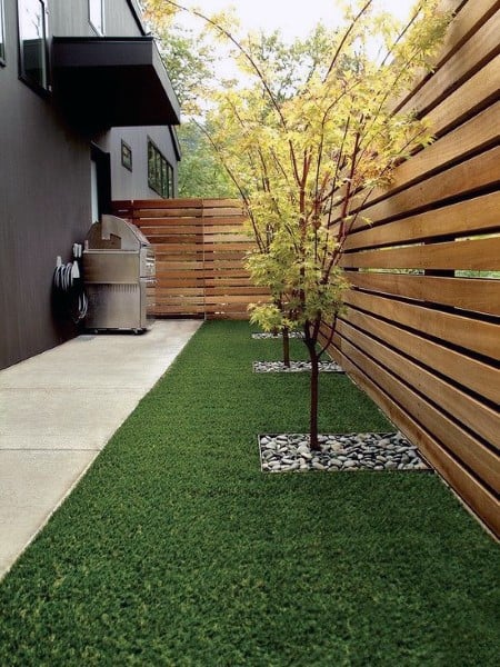 Contemporary horizontal wooden fence lining a narrow backyard with artificial grass and decorative trees.
