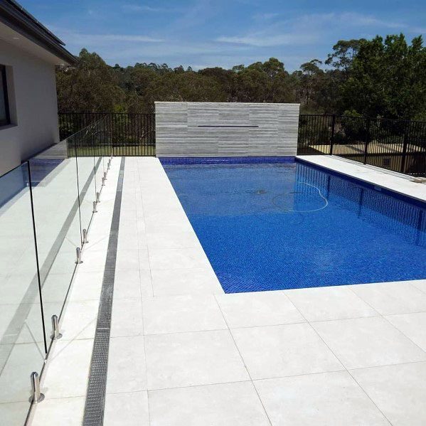Modern pool with deep blue mosaic tiles, glass fencing, and a spacious white stone deck.