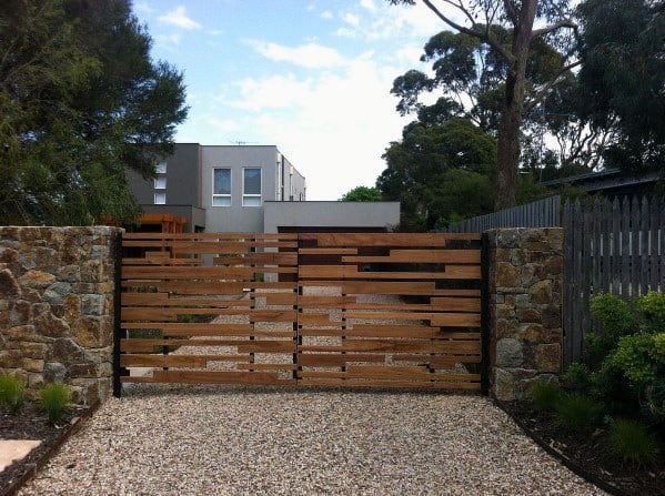 A modern wooden gate with stone pillars leads to a contemporary house, surrounded by trees and a gravel pathway