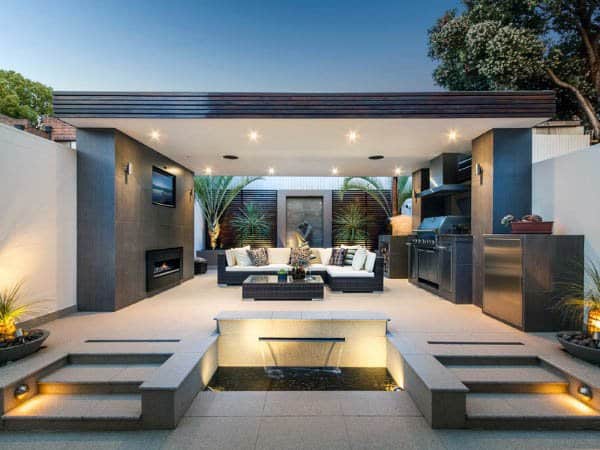 Modern outdoor patio featuring seating, a fireplace, and grill area, surrounded by plants and illuminated by spotlights