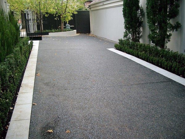 Textured asphalt driveway with white stone edging bordered by neatly trimmed hedges and trees.