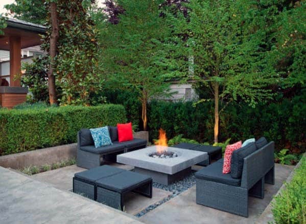 Outdoor seating area with black sofas, colorful pillows, and a square fire pit surrounded by lush green trees