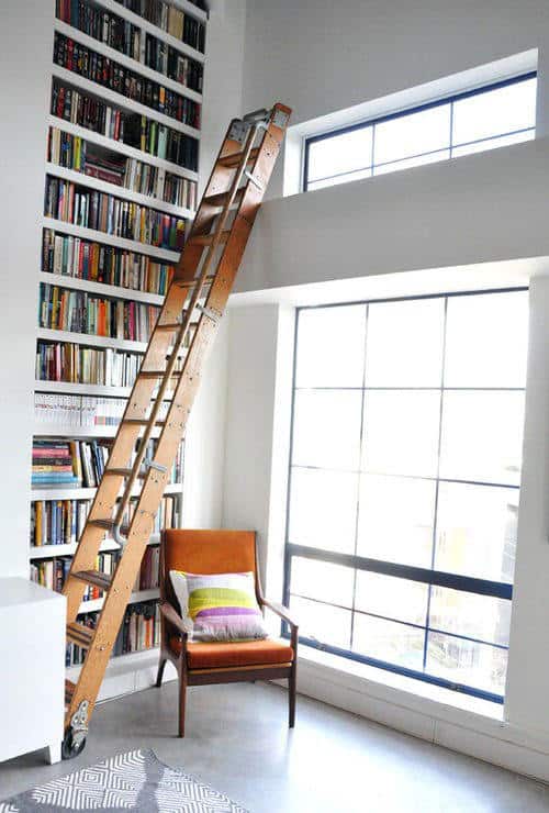 Tall bookshelf with a wooden ladder, like a cozy library corner, beside a chair with colorful pillows by large windows