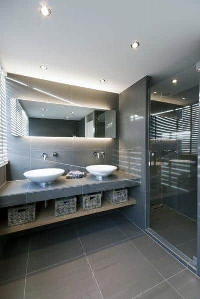 A modern bathroom with gray tiles, a double vanity, under-sink baskets, and a glass-enclosed shower.