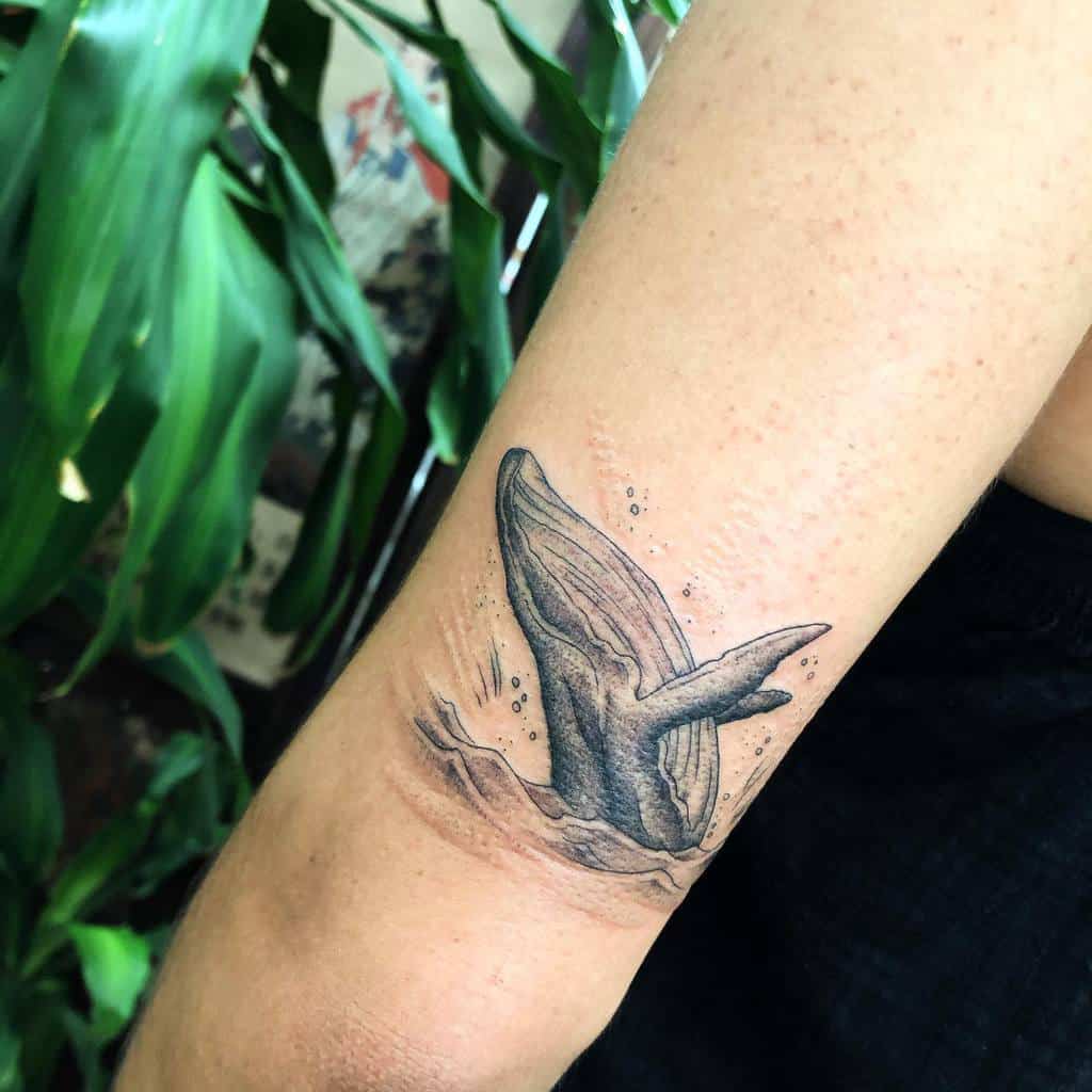 Black and gray tattoo of a breaching whale with water ripples and bubbles from ocean