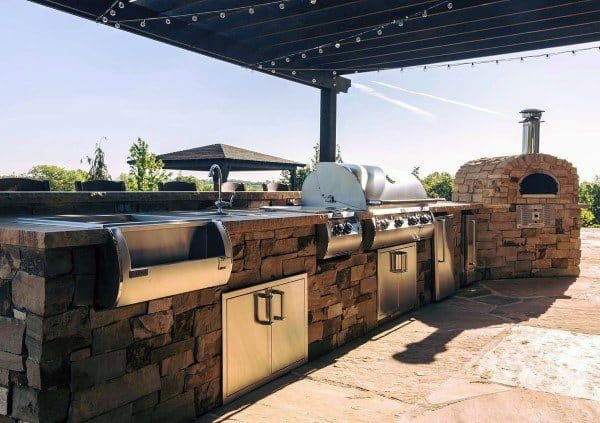 Outdoor kitchen with brick counter, stainless grill, sink, and pizza oven under a pergola—all set in an outdoor oasis