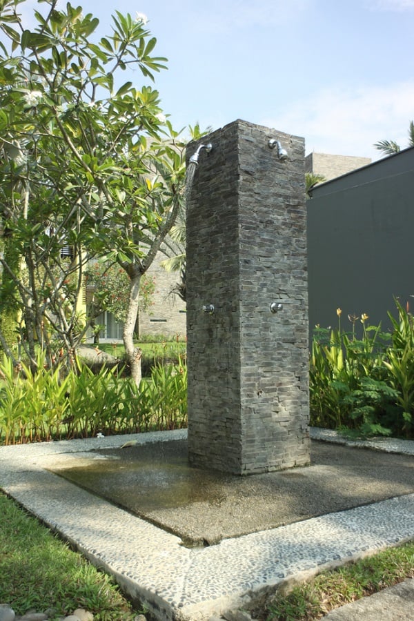 Freestanding outdoor shower with stone pillar design, surrounded by lush greenery and pebble flooring.