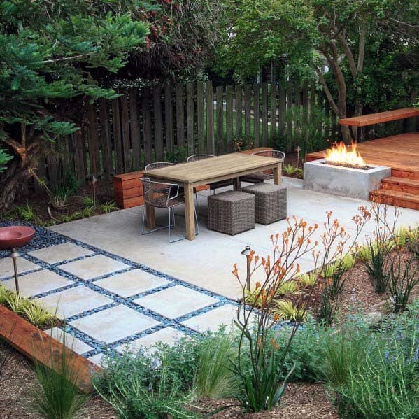 Modern patio with a dining table, comfortable seating, and a firepit, featuring a river rock-bordered pathway and lush greenery