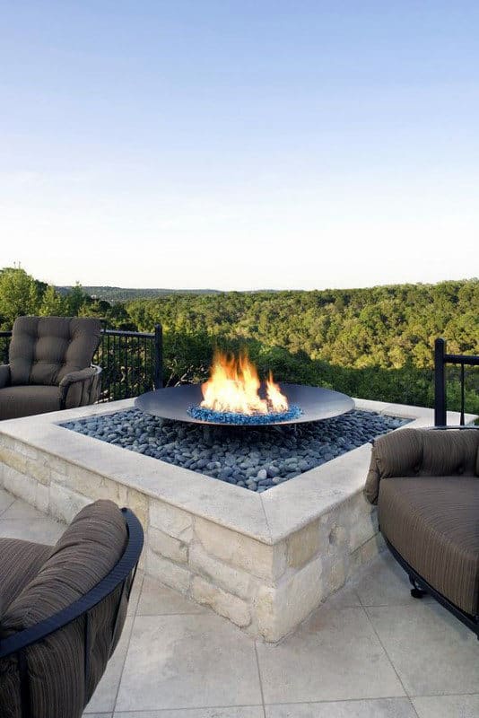 Fire pit with blue glass stones, surrounded by cushioned chairs, overlooking a lush green landscape