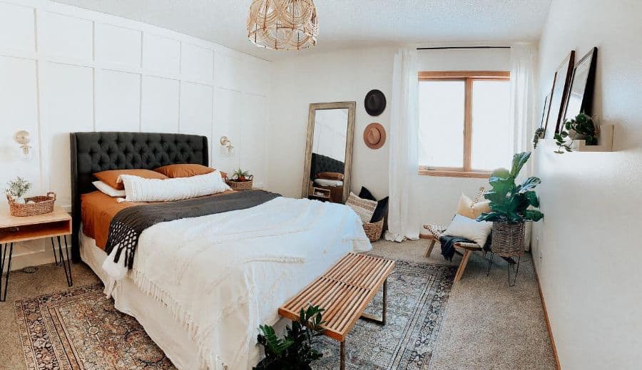 A cozy bedroom featuring a large white bed, plants, a bench, and a window, with decor including a mirror and wall art