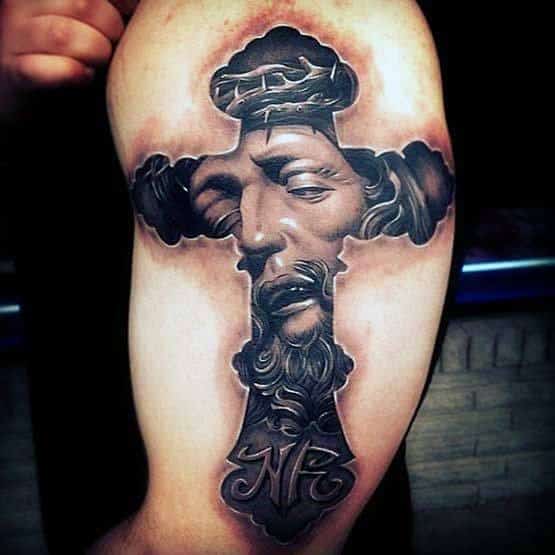 Tattoo of a detailed cross with a somber face inside on a person's upper arm