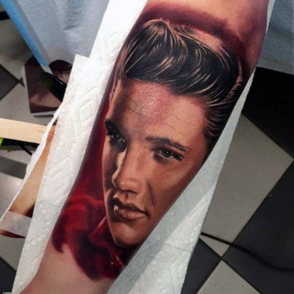 Realistic color tattoo of Elvis Presley on forearm, featuring smooth shading, a red-toned background, and his iconic hairstyle.