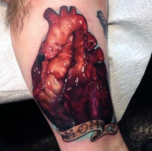 Hyper-realistic anatomical heart tattoo with rich red and brown tones, placed above a scroll banner with lettering.