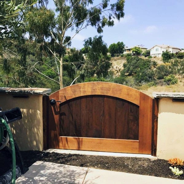 Wooden gate in a garden, surrounded by trees, overlooking a hillside with distant houses under a clear sky