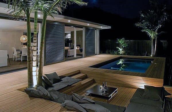 Luxury wooden pool deck with lounge seating, palm trees, and ambient night lighting.