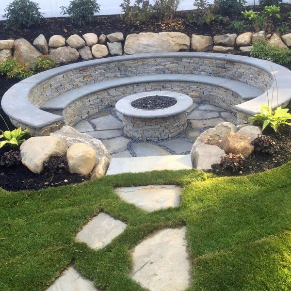 Stone sunken fire pit area with circular seating, surrounded by landscaping and greenery.