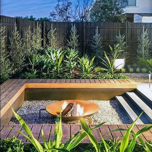 Wooden deck with a metal fire bowl, gravel area, and lush greenery in a modern backyard.