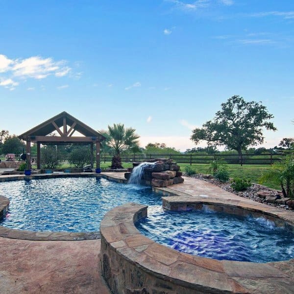 Backyard pool with rock waterfall, spa, and wooden gazebo surrounded by lush greenery.