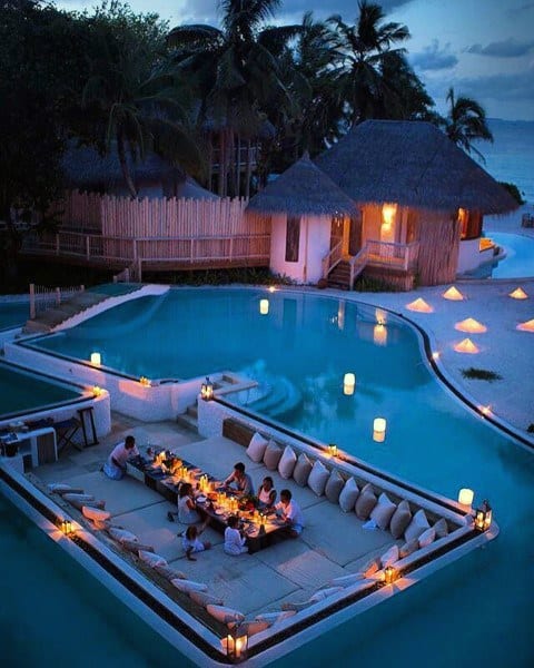 Luxurious poolside dining area with cushions, lanterns, and tropical thatched huts.