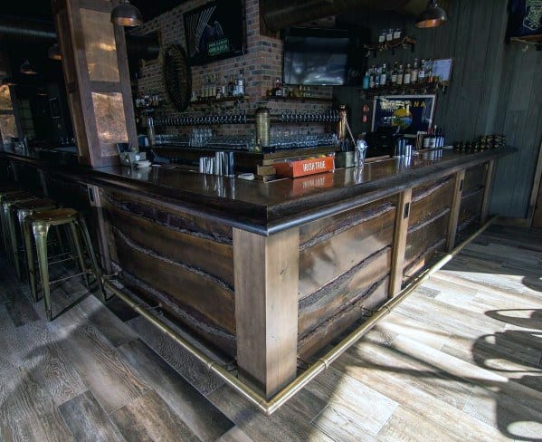 Rustic bar with brass foot rail, wooden bar panels, and industrial-style metal stools on wood flooring.