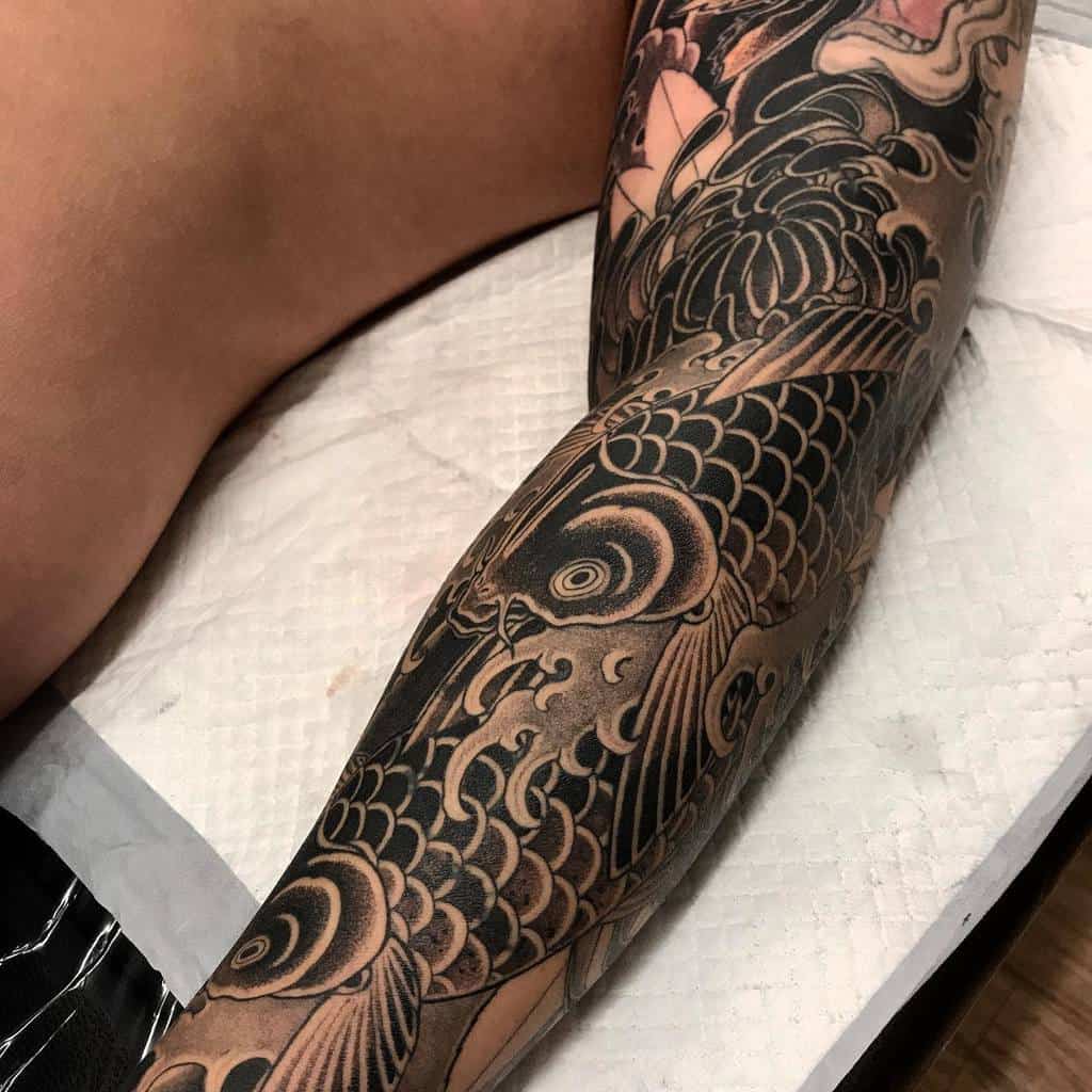 Detailed black and gray koi fish tattoo covering a person's leg, featuring water and floral motifs