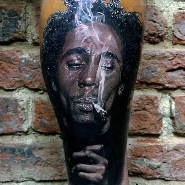 Realistic tattoo of Bob Marley with eyes closed, smoking, and a trail of smoke rising, done in full color on the lower leg.